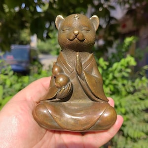 Puede incluir: Figura de bronce de un oso en posición de meditación sentado. El oso lleva una túnica y tiene las manos juntas en oración. Sostiene dos pequeñas calabazas. La figura mide aproximadamente 10 cm de altura.