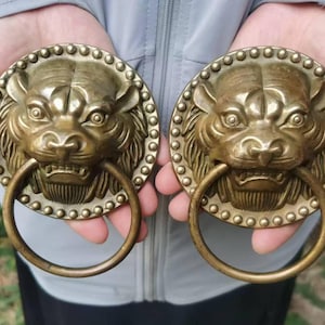 Op de afbeelding: Een paar ronde deurkloppers van messing met een gedetailleerd reliëf van een tijgerkop. Elk exemplaar is gemonteerd op een ronde plaat met een decoratieve kralenrand en is voorzien van een grote, gladde, zwaaiende ring aan de bek.