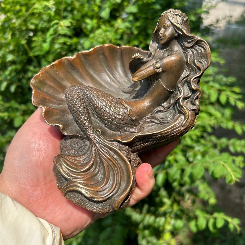 May include: A bronze-toned sculpture of a mermaid resting inside a large, open seashell. The mermaid has long, flowing hair and a detailed tail, while the shell features prominent ridges. The entire piece has a metallic finish with dark highlights.