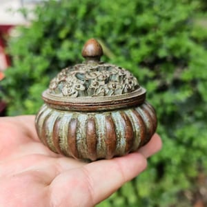 May include: A small, antique bronze container with a lid. The container has a rounded, ribbed body and a lid adorned with floral patterns. The lid is topped with a small, conical finial. The bronze has a weathered, aged appearance with hints of green.