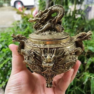 May include: An ornate brass incense burner with dragon designs. The lid features a dragon sculpture, and the body is decorated with detailed dragon carvings. The burner has three feet and dragon head handles. The colour is a warm, antique gold.
