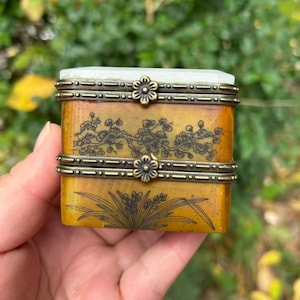 May include: A rectangular, antique-style box with a light green top and a yellow-brown body. The box features floral designs and decorative metal accents. The box is held in a hand.