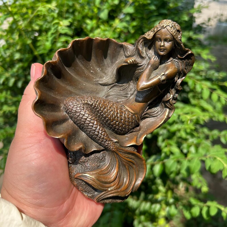 May include: A bronze-toned decorative figurine featuring a mermaid resting inside a large, open seashell. The mermaid has long, flowing hair and a detailed scaly tail, while the shell forms a curved, bowl-like structure behind her figure.