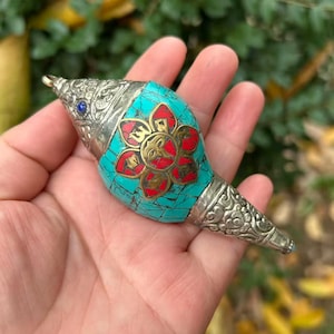 Peut inclure: Un coquillage décoratif avec une mosaïque turquoise, des accents floraux rouges et des détails en métal argenté. La coquille présente une petite pierre précieuse bleue et des motifs complexes. Le coquillage est tenu dans une main.