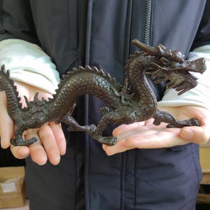 以下が含まれることがあります： 精巧なディテールが施されたブロンズ製のドラゴン彫刻。 ドラゴンは、体をアーチ状にし、足を伸ばしたダイナミックなポーズで描かれています。 彫刻は質感のある表面と濃い茶色をしています。 ドラゴンの頭には角と開いた口が詳細に表現されています。