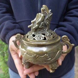 Puede incluir: Un quemador de incienso de color bronce con una figura de dragón en la parte superior. El quemador tiene intrincados patrones florales y de nubes, con asas y patas ornamentadas. El diseño general es detallado y de aspecto antiguo.