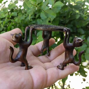Könnte beinhalten: Eine bronzefarbene Katzenfigur, die ein kleines, rundes, gemustertes Tablett hält. Drei Katzenfiguren stützen das Tablett mit ihren Armen. Die Katzen stehen mit eingerollten Schwänzen.
