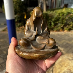 May include: A bronze-toned figurine of a hooded figure in a seated meditation pose with hands pressed together in a prayer position. The sculpture features smooth, flowing lines and a rounded base, capturing a serene and contemplative form.