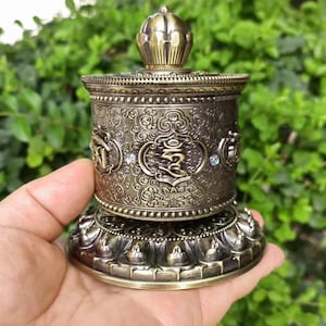 Puede incluir: Una antigua rueda de oración de bronce con intrincados patrones en relieve y detalles decorativos. La rueda presenta detalles ornamentados, incluyendo símbolos religiosos y pequeños adornos brillantes. La base y la parte superior están adornadas con diseños florales y geométricos.