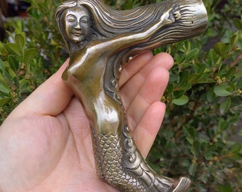 May include: Bronze-coloured mermaid-shaped handle. The mermaid has flowing hair and a scaled tail. The handle is curved and appears to be made of metal. The mermaid is in a relaxed pose, with her arm outstretched.