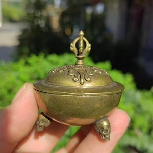 Puede incluir: Un pequeño quemador de incienso antiguo de latón con una tapa decorativa y patas en forma de calavera. La tapa presenta detalles ornamentados y un remate estilizado. El quemador se sostiene en una mano, mostrando su tamaño compacto y su intrincado diseño.