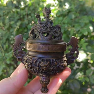 May include: A bronze incense burner with intricate dragon and mythical creature detailing. The burner features a lid with a dragon sculpture and ornate handles. The dark brown color and detailed carvings give it an antique appearance.