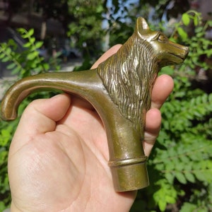 May include: A bronze-coloured cane handle with a detailed wolf head design. The handle curves smoothly, with the wolf's head at the top. The wolf has detailed fur and a focused expression. The handle is held in a hand.