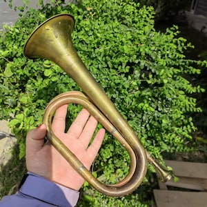 May include: A vintage brass bugle with a flared bell and curved tubing. The instrument has a tarnished, aged appearance, suggesting it is antique. The bugle is held against a backdrop of green foliage.