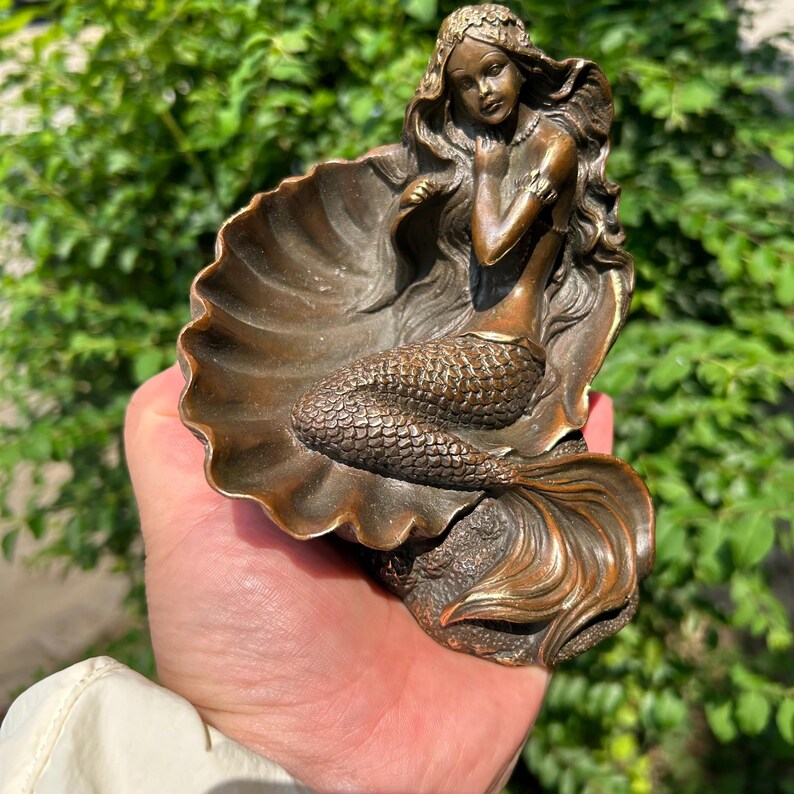 May include: A bronze-toned decorative figurine featuring a mermaid with long flowing hair resting inside a large, open seashell. The mermaid has a detailed scaly tail and is positioned with one hand near her face, set against a textured base.