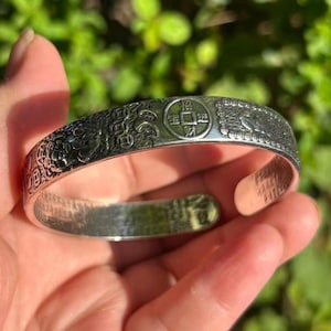 May include: Silver cuff bracelet with engraved Chinese characters and symbols. The open cuff design features detailed relief patterns, including a coin and bird motifs. The bracelet is held in a hand against a blurred green background.