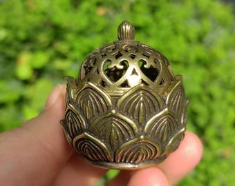 May include: A small, antique brass incense burner. The burner is shaped like a lotus flower with intricate cut-out designs on the lid. The overall color is a warm, aged brass, and the details are finely crafted. The burner is held in a hand.