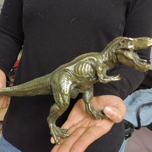 May include: A bronze-colored Tyrannosaurus Rex figurine. The dinosaur is depicted in a dynamic pose with its mouth open, showcasing detailed texture and musculature. The figurine is held in a person's hands, highlighting its size and craftsmanship.