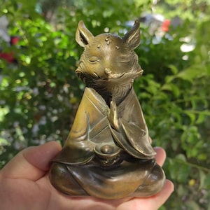 May include: A bronze fox statue in a meditative pose, with closed eyes and hands clasped in prayer. The fox is wearing a robe and has a serene expression. The statue is approximately 4 inches tall.