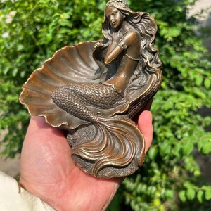 May include: A bronze-toned decorative figurine featuring a mermaid with long flowing hair, seated within a large, open seashell. The mermaid has a detailed scaly tail and is positioned against the curved, ribbed interior of the shell structure.