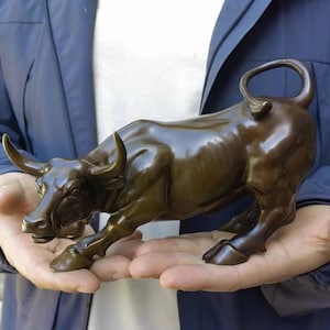 Op de afbeelding: Een bronzen sculptuur van een stier in een aanvallende houding. De stier heeft gebogen hoorns en een gespierde bouw, met een gepolijst, reflecterend oppervlak. De sculptuur wordt in open handen gehouden, waarbij de details en vorm worden getoond.