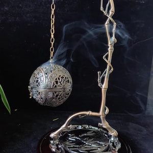 Op de afbeelding: Een sierlijke, bronskleurige wierookbrander met een bolvormige censer in filigraanstijl, opgehangen aan een bamboe-achtige standaard. Wispelturige rook stijgt op uit de censer, wat gebruik suggereert. De basis is donker hout met een decoratieve metalen plaat.