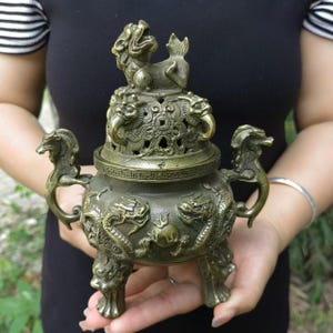 Puede incluir: Quemador de incienso de bronce con intrincados detalles de dragones y criaturas míticas. El quemador presenta una tapa con una pequeña estatua en la parte superior y asas a los lados. El cuerpo del quemador está adornado con motivos de dragones y descansa sobre tres pies.