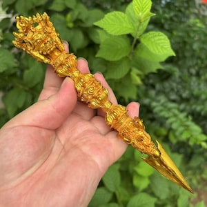 May include: A detailed, gold-coloured ritual dagger with ornate carvings. The dagger features a pointed blade and a handle adorned with figures and intricate patterns. The design suggests a ceremonial or religious artifact.