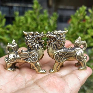 May include: Two brass-colored, mythical lion statues with wings and detailed carvings. The statues face each other, showcasing open mouths and intricate mane details. The background is blurred green foliage.