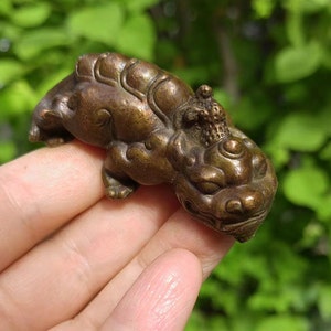 May include: A bronze-colored figurine of a mythical creature, possibly a Foo Dog or similar guardian figure. The sculpture has detailed features, including a lion-like face and ornate detailing on its back. The figure is held in a hand against a blurred green background.