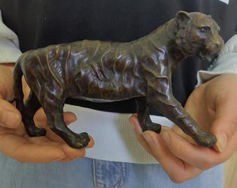 May include: A bronze-colored tiger figurine with detailed fur texture. The sculpture depicts the tiger in a walking pose, showcasing its muscular build and facial features. The figurine is held in open hands.