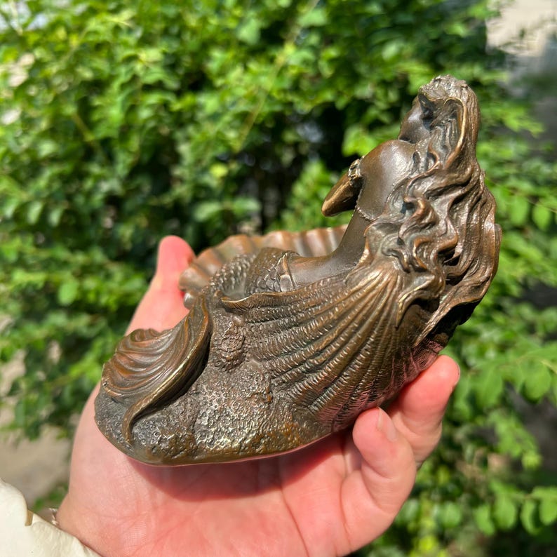 May include: A bronze-toned sculpture of a mermaid resting within a large, textured seashell. The figure features long, flowing hair and a detailed tail, with the entire piece showcasing a metallic finish and intricate, raised relief patterns.