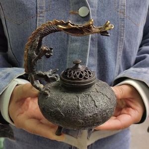 Puede incluir: Un quemador de incienso de metal en tono bronce oscuro con un cuerpo redondeado y texturizado cubierto de caracteres en relieve. El asa tiene forma de dragón detallado con el cuerpo curvado y la tapa presenta un patrón calado decorativo.