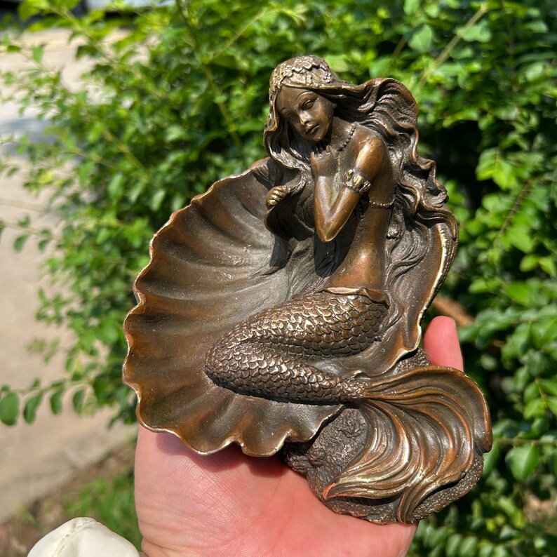 May include: A bronze-toned mermaid figurine resting inside a large, open seashell. The mermaid has long, flowing hair, a detailed scaled tail, and is positioned with one hand near her neck. The sculpture features a metallic finish with dark highlights.