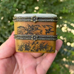 Puede incluir: Una caja rectangular de estilo antiguo con tapa con bisagras. La caja es de color ámbar cálido con intrincados diseños negros grabados de pájaros, peces y follaje. Los bordes están adornados con una banda metálica decorativa y detalles florales.