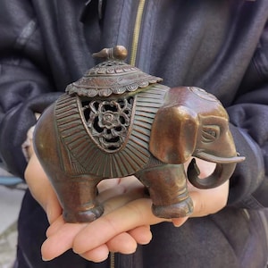 以下が含まれることがあります： 精巧な花柄と幾何学模様が施されたブロンズ製の象の置物。象は装飾的な鞍と背中に小さな蓋が付いています。この置物は手で持たれており、その詳細な職人技とアンティークな外観を示しています。