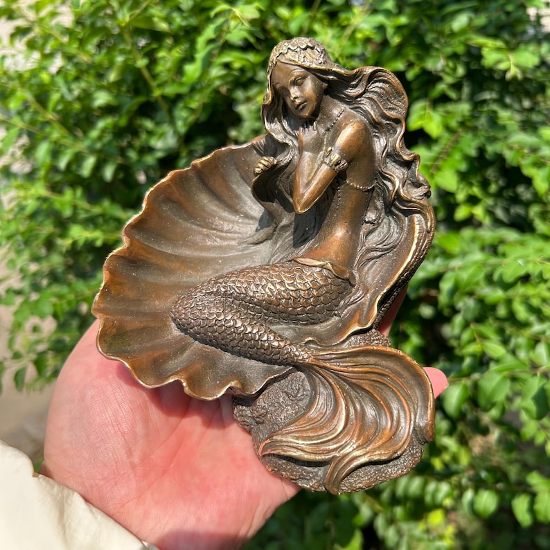 May include: A bronze-toned decorative dish shaped like a large scallop shell featuring a sculpted mermaid figure resting inside. The mermaid has long flowing hair and a detailed scaled tail, with the entire piece finished in a metallic brown.