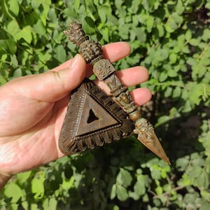 May include: An antique bronze ritual dagger with a triangular base and detailed carvings. The dagger has a sharp, pointed blade and a decorative handle. The base features a central triangular aperture. The background is green foliage.