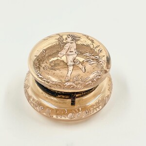 May include: A clear, antique-style glass jar lid with a detailed relief of a child running through a field. The lid is a light amber color, and the base has a decorative floral pattern. The jar lid is likely for a vintage apothecary or kitchen storage.