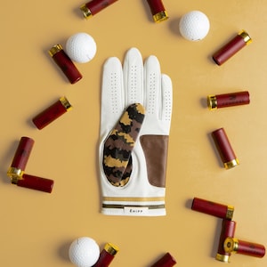 Puede incluir: Un guante de golf blanco con un patrón de camuflaje y detalles en cuero marrón, rodeado de pelotas de golf y cartuchos de escopeta rojos con casquillos dorados. El guante tiene la marca "E HIPP" en la muñeca.