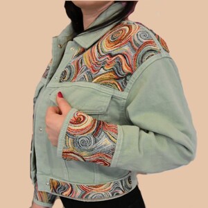 May include: A light green denim jacket with colorful embroidered swirl patterns on the shoulders, cuffs, and hem. The jacket has a classic design with button closures and two chest pockets. The embroidery features shades of red, yellow, orange, and blue.