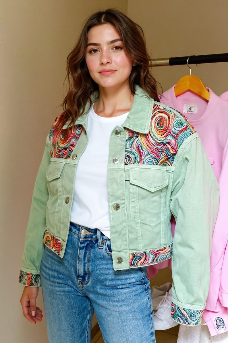 May include: A light green denim jacket with colorful embroidered details on the shoulders, cuffs, and hem. The jacket has a classic design with button closures and pockets. The model is wearing a white t-shirt and blue jeans.