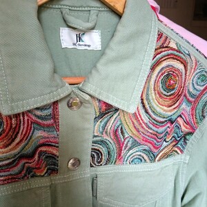 May include: A light green jacket with a colorful, swirling pattern on the upper chest. The jacket has silver buttons and a small label that reads "HC-Revamp". The jacket is hanging on a wooden hanger.