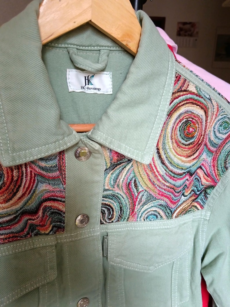 May include: A light green jacket with a colorful, swirling pattern on the upper chest. The jacket has silver buttons and a small label that reads "HC-Revamp". The jacket is hanging on a wooden hanger.