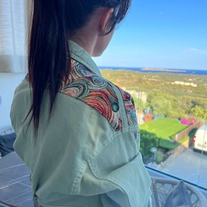May include: A light green denim jacket with colorful embroidered shoulder and waist details. The jacket has a classic design with a button-up front and pockets. The embroidery features swirls of red, blue, and gold. The jacket is a stylish outerwear piece.