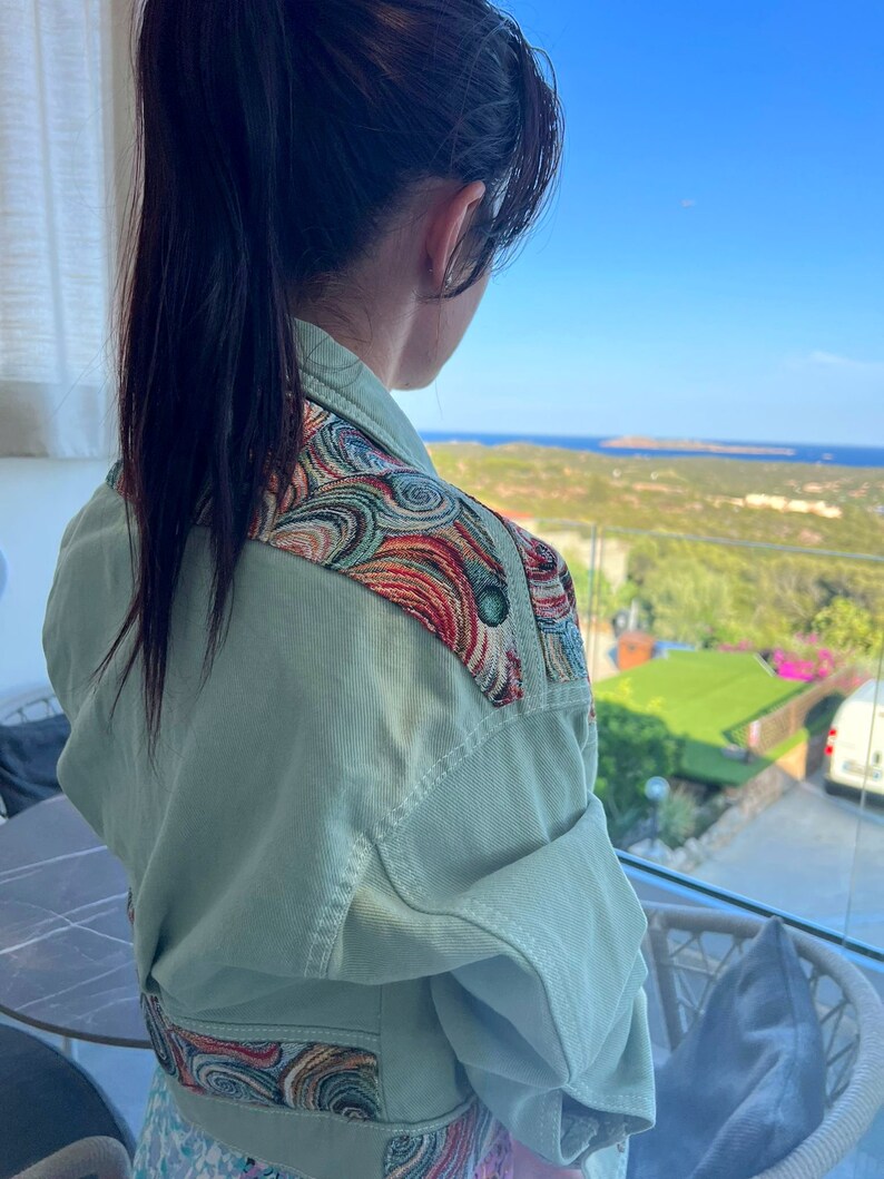 May include: A light green denim jacket with colorful embroidered shoulder and waist details. The jacket has a classic design with a button-up front and pockets. The embroidery features swirls of red, blue, and gold. The jacket is a stylish outerwear piece.