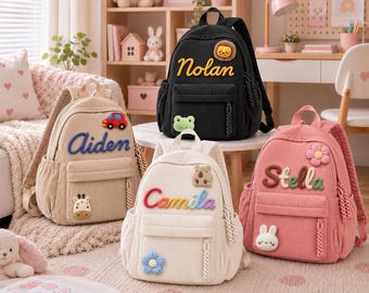 Puede incluir: Cuatro mochilas de pana en beige, negro, blanco y rosa. Cada mochila presenta un nombre en letras grandes, coloridas y texturizadas, junto con pequeños apliques de peluche como un coche, león, rana, oso, flor, jirafa y conejo.