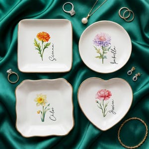 May include: Four white ceramic ring dishes with gold rims, each featuring a different floral design and a name. The flowers include an orange marigold, a purple thistle, a yellow daffodil, and a pink carnation. The names are Sienna, Isabella, Chloe, and Jessica.
