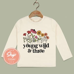 A white long-sleeved t-shirt with a floral design and the text "young wild & three" printed on it, hanging on a wooden hanger.