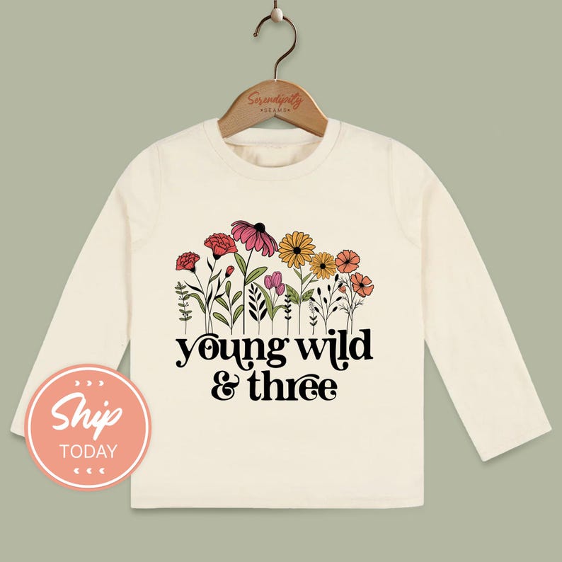 A white long-sleeved t-shirt with a floral design and the text "young wild & three" printed on it, hanging on a wooden hanger.